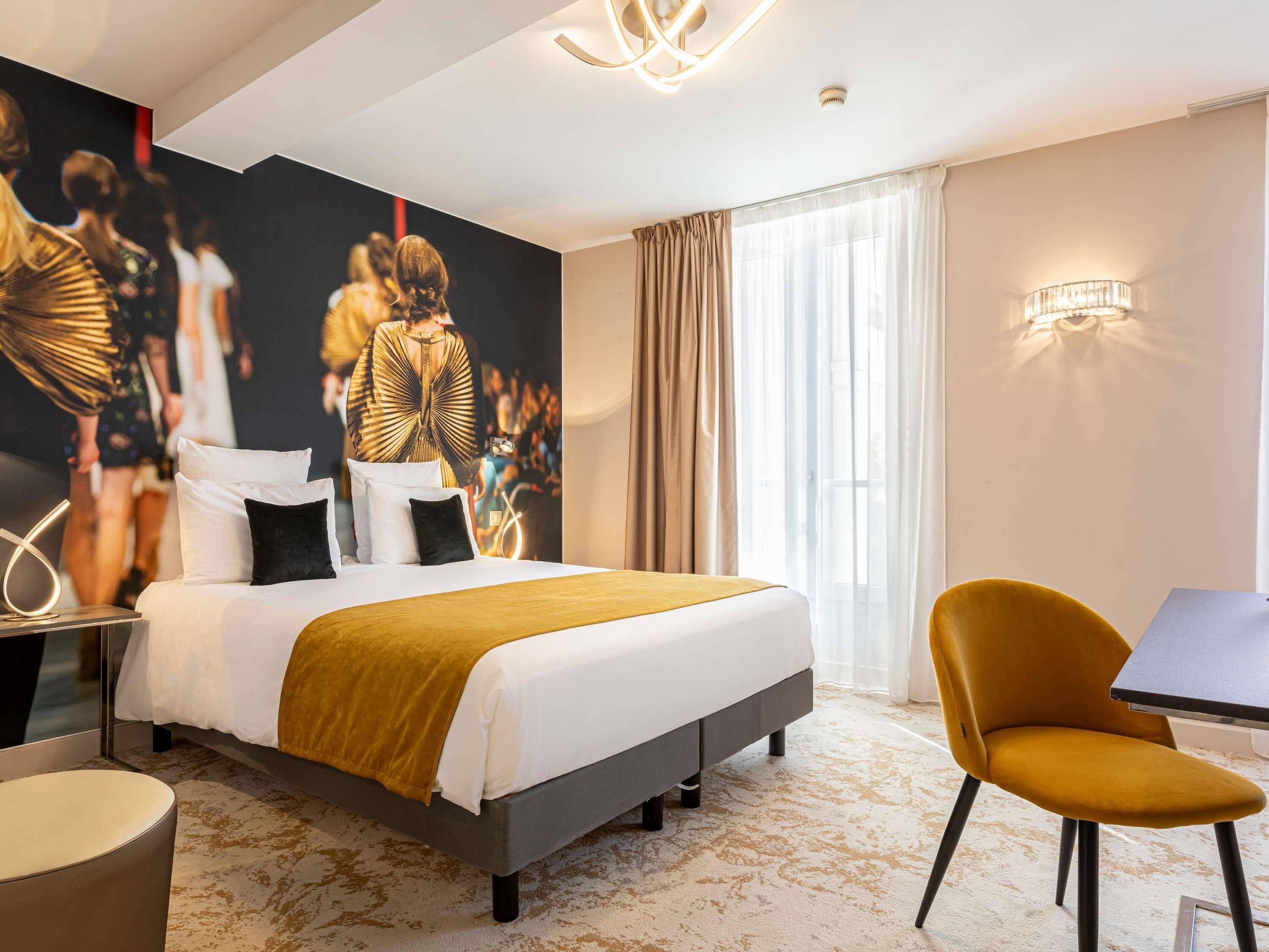 Ein Hotelzimmer mit Bett, Sessel, Tisch, einer Wand mit einem Poster und einem Vorhangfenster.