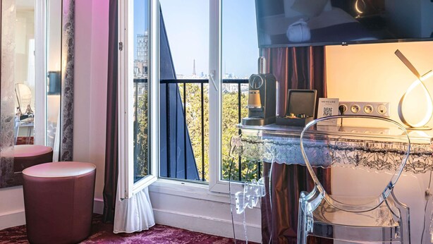 Quarto de hotel moderno com uma janela grande, televisão de tela plana, máquina de café e mesa de vidro com toalha de renda.