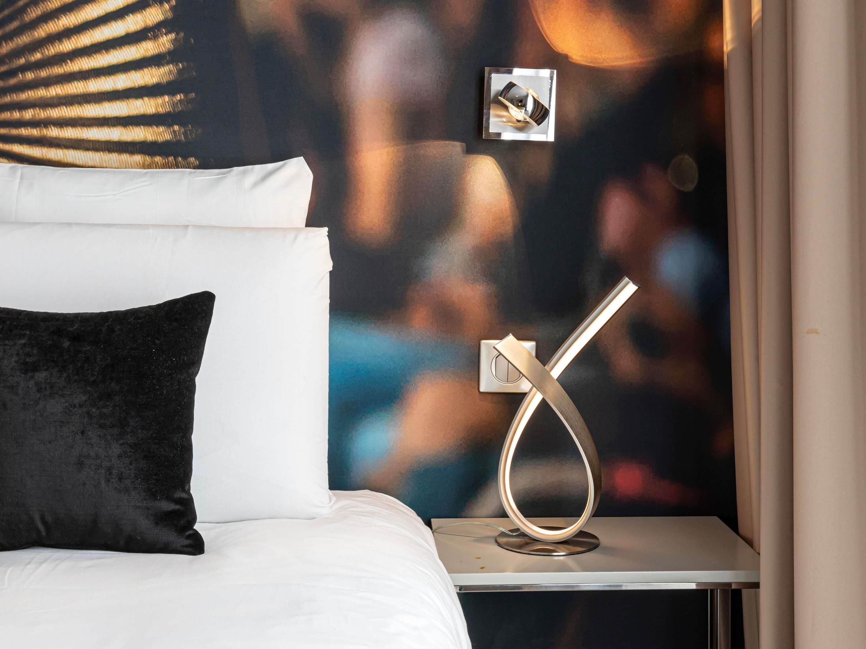 Una cama con almohadas blancas y negras, una mesita de noche con una lámpara moderna y una pared con fondo desenfocado.