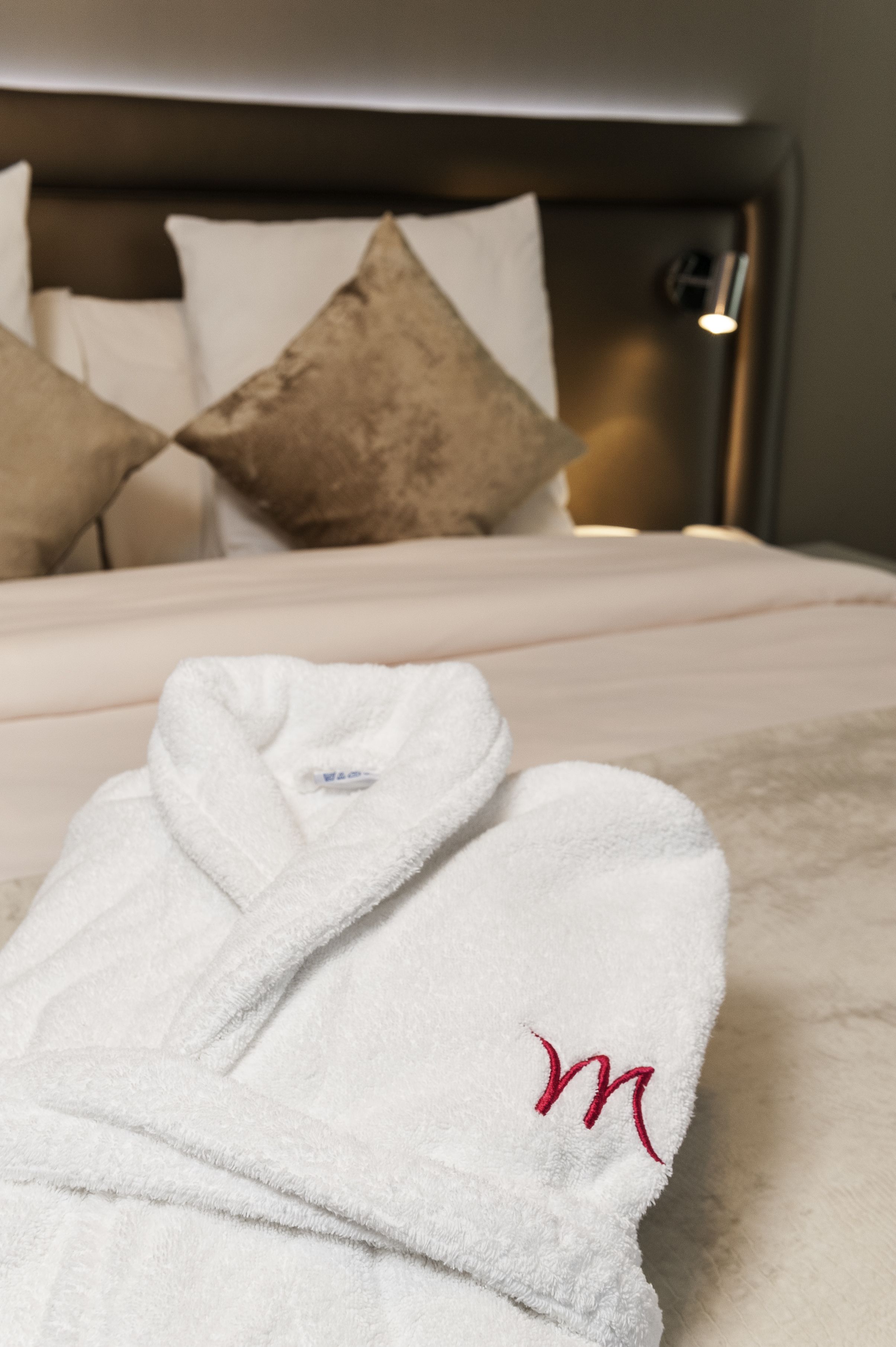 A white bathrobe with a red 'M' logo on a bed with a headboard and pillows.