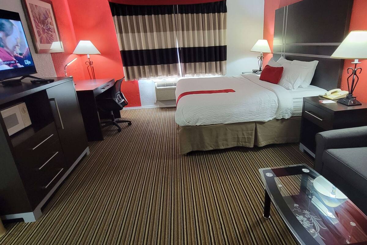A hotel room with a bed, bedside tables with lamps, a desk with a TV, a sofa, and a carpeted floor.