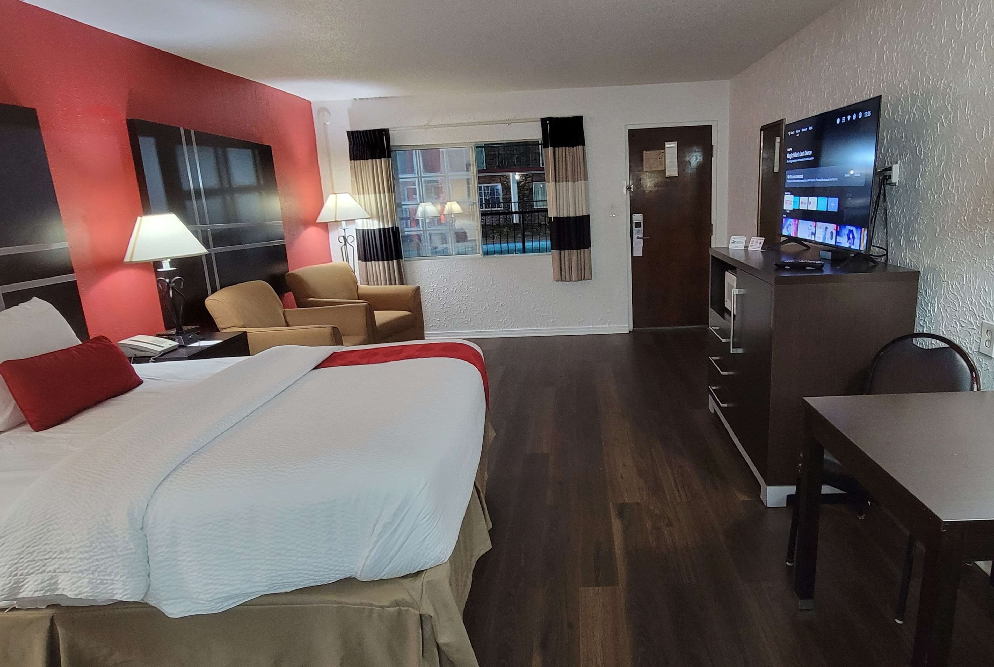 A hotel room with a bed, two chairs, a desk, a television, and a window with curtains.