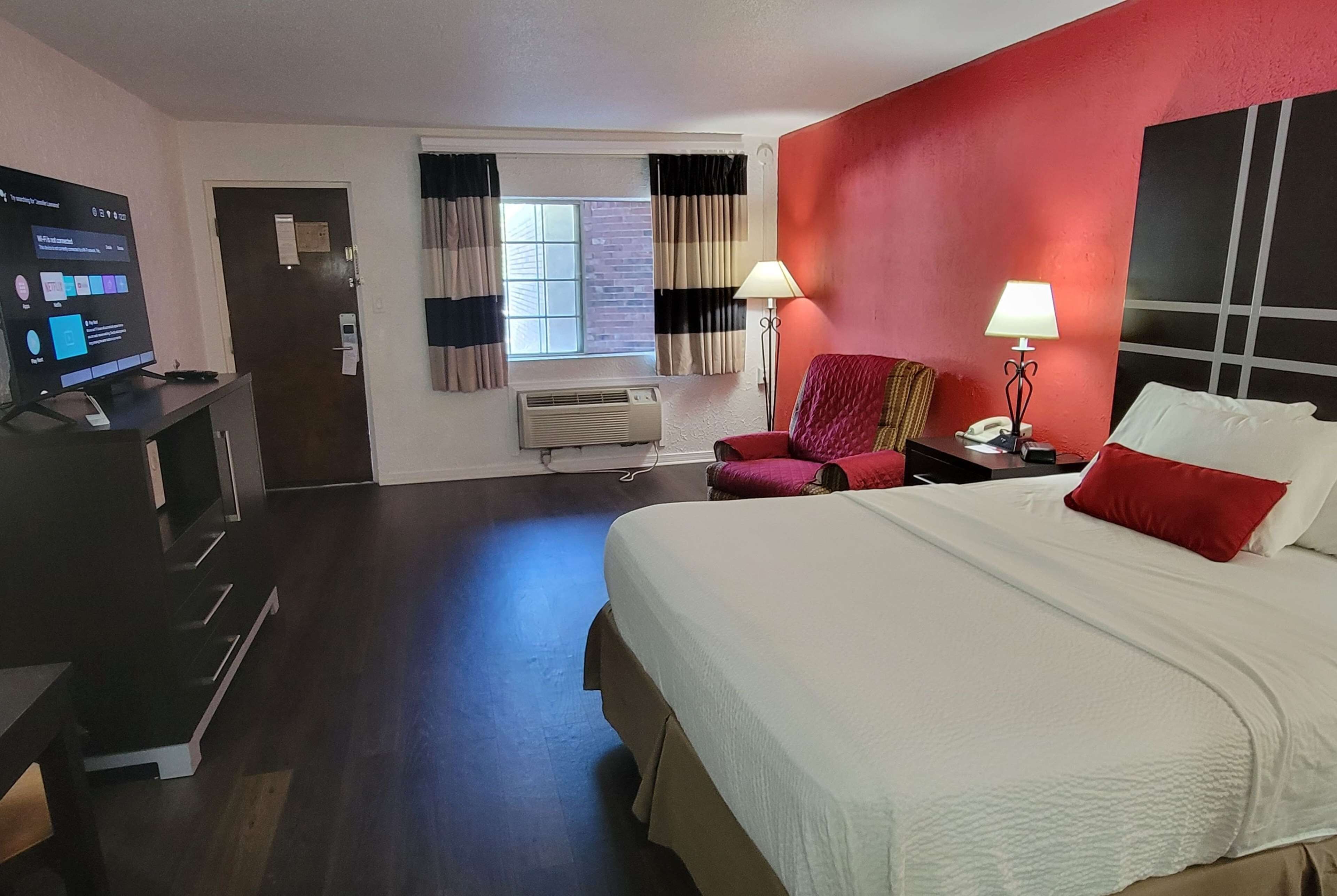 Hotelkamer met een bed, een bureau met een computer, een stoel, een raam met gordijnen en een rode accentmuur.