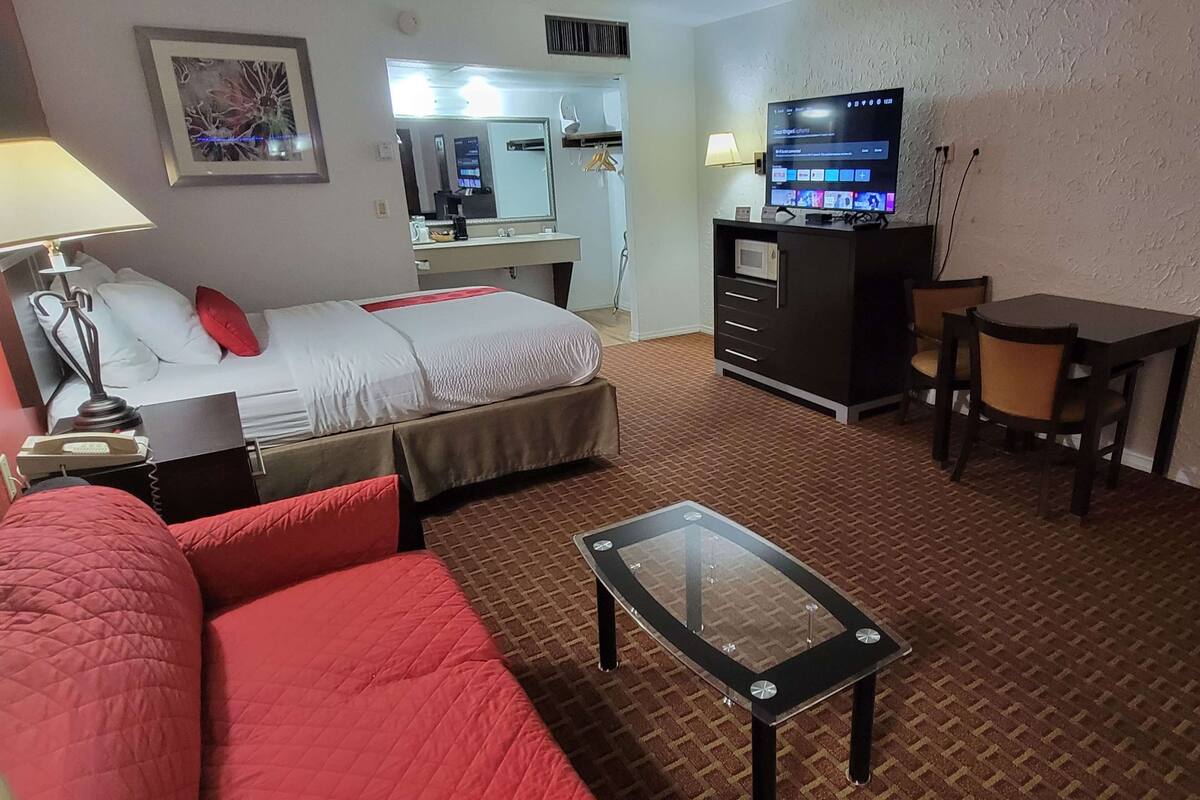 A hotel room with a bed, a red couch, a glass-top table, a desk with a TV, a mirror, and a wardrobe.