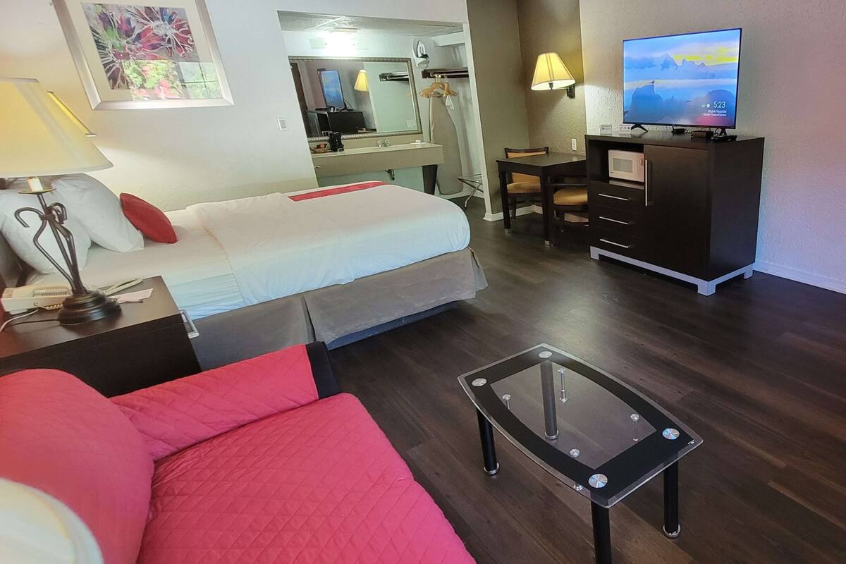 A hotel room with a bed, a red sofa, a glass coffee table, a desk with a chair, a TV, and a wardrobe.