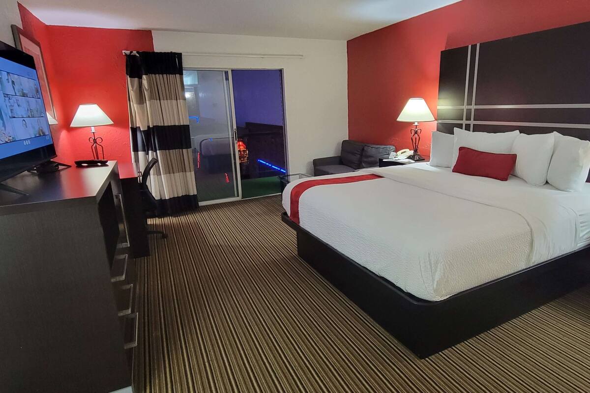 A hotel room with a large bed, two bedside tables with lamps, a desk with a computer, a sofa, and a sliding glass door leading to a balcony.