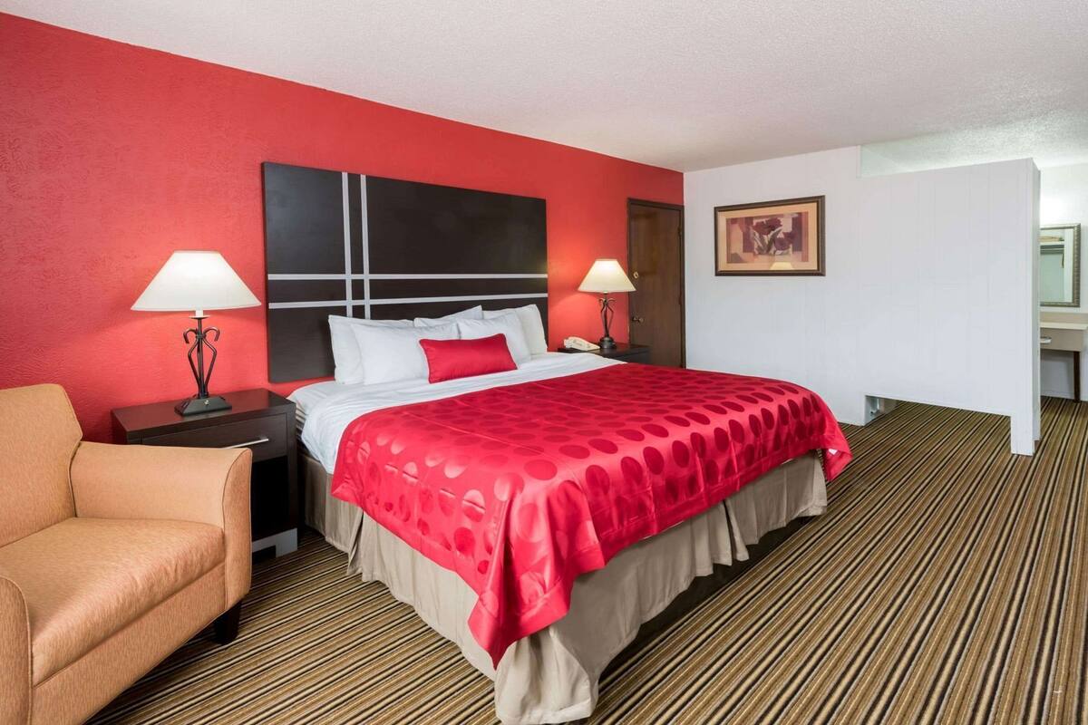 A hotel room with a bed, two bedside tables with lamps, a chair, a red wall, and a striped carpet.