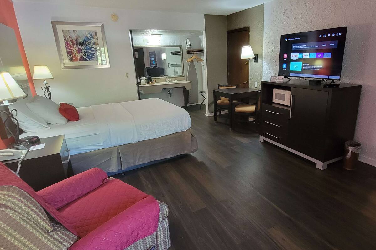 A hotel room with a bed, a desk, a chair, a TV, and a bathroom visible through an open door.
