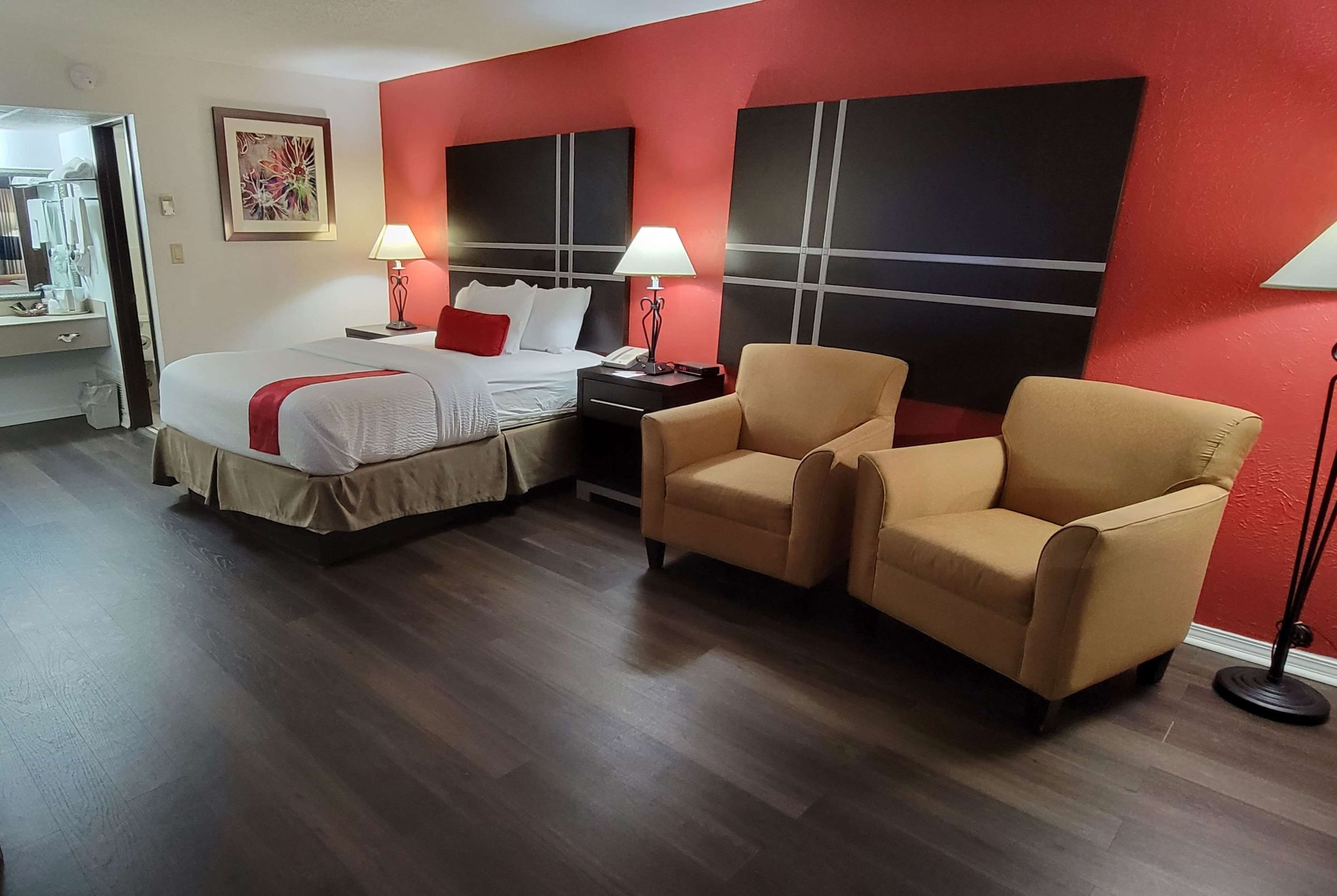 Quarto de hotel com uma cama grande, duas poltronas, uma mesa de cabeceira com abajur e uma parede de destaque vermelha.