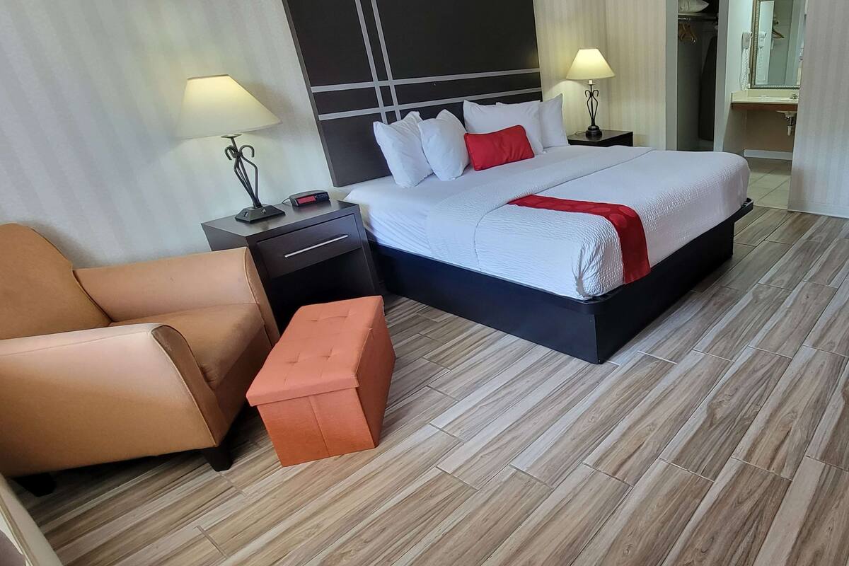A hotel room with a bed, a nightstand, a lamp, a chair, and a small ottoman.