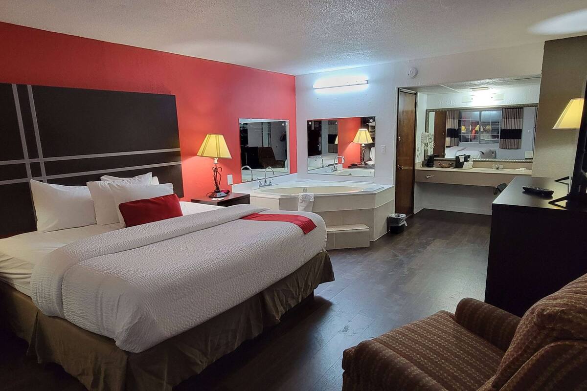 A hotel room with a large bed, a bedside table, a mirror, a desk with a lamp, and a bathroom with a sink and mirror.
