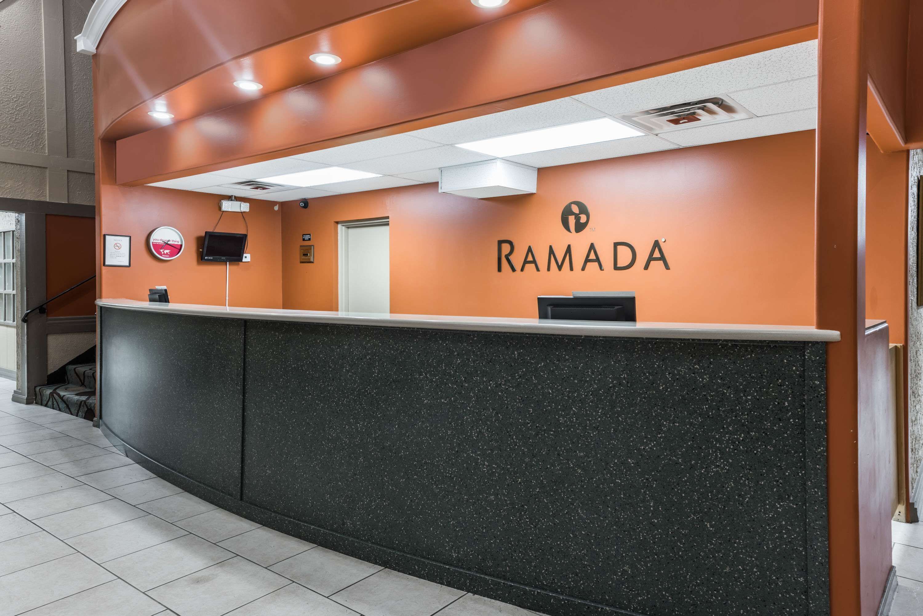A reception desk with a black granite top and orange walls, featuring the name 'RAMADA' prominently displayed.
