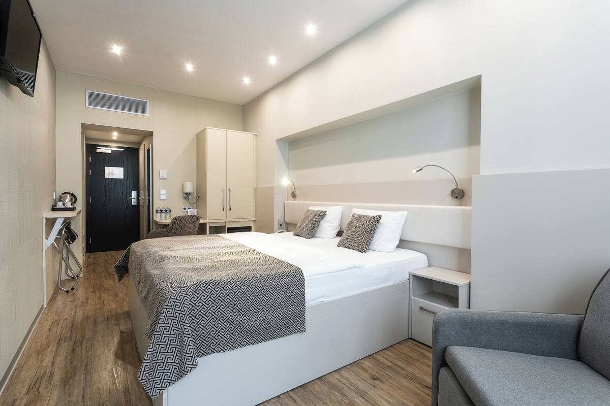 A modern hotel room with a large bed, bedside tables, a chair, and a wardrobe.