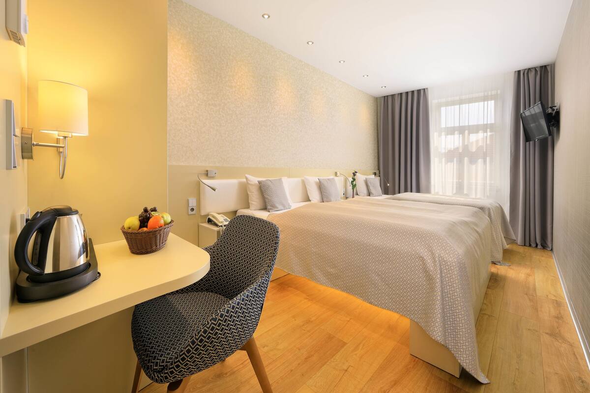 A hotel room with a bed, a desk with a kettle, a chair, and a TV mounted on the wall.