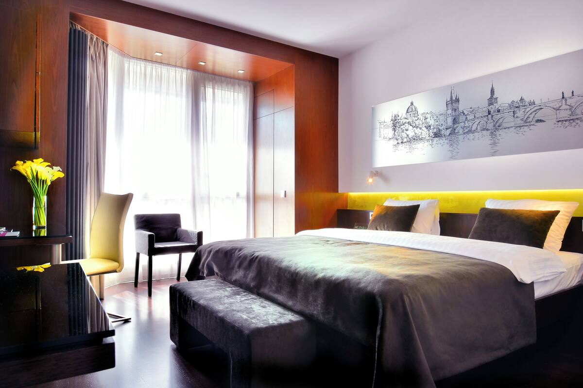 A modern hotel room with a large bed, a bedside table, a chair, and a view of a cityscape.