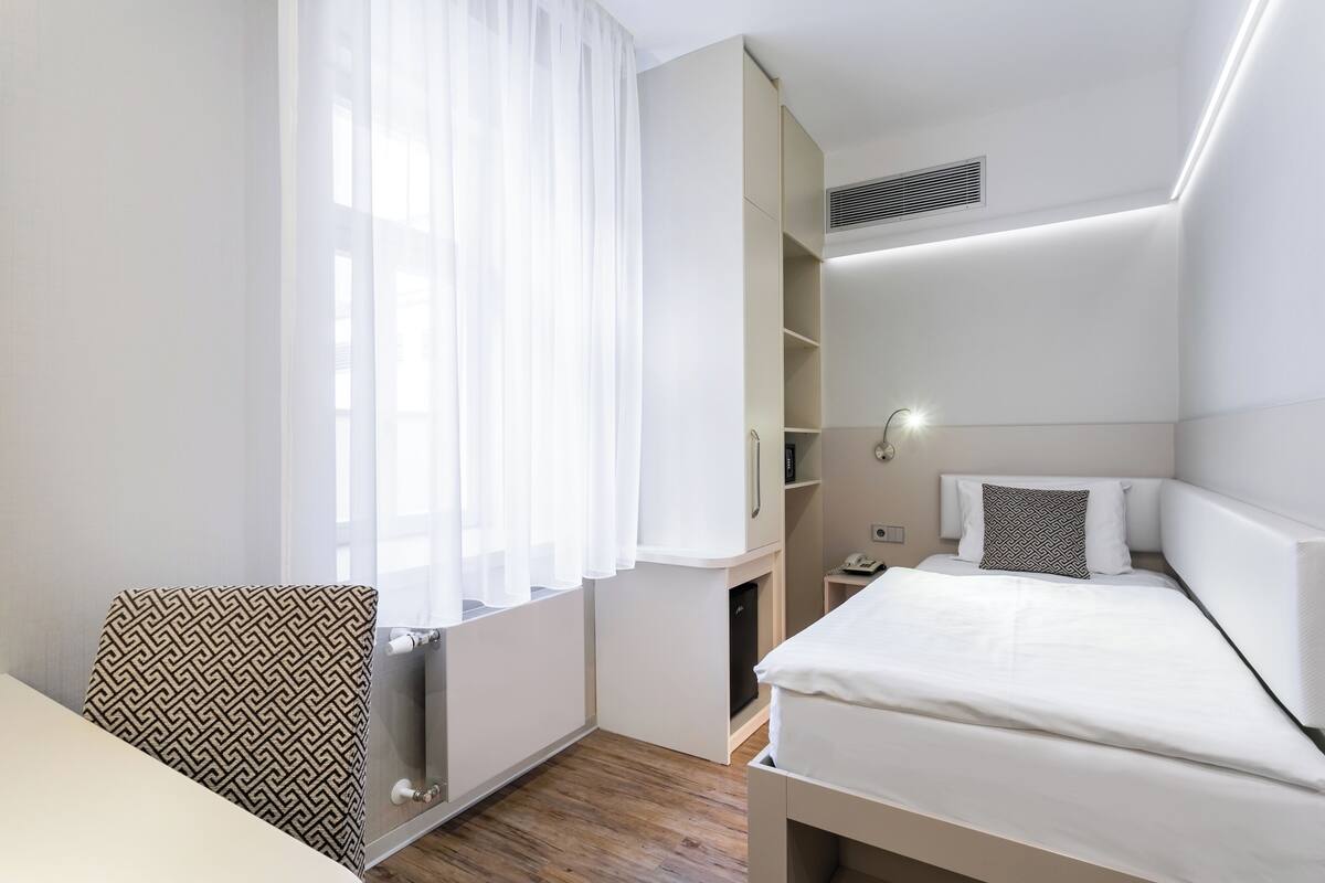 A compact hotel room with a bed, a chair, a wardrobe, and a window with sheer curtains.