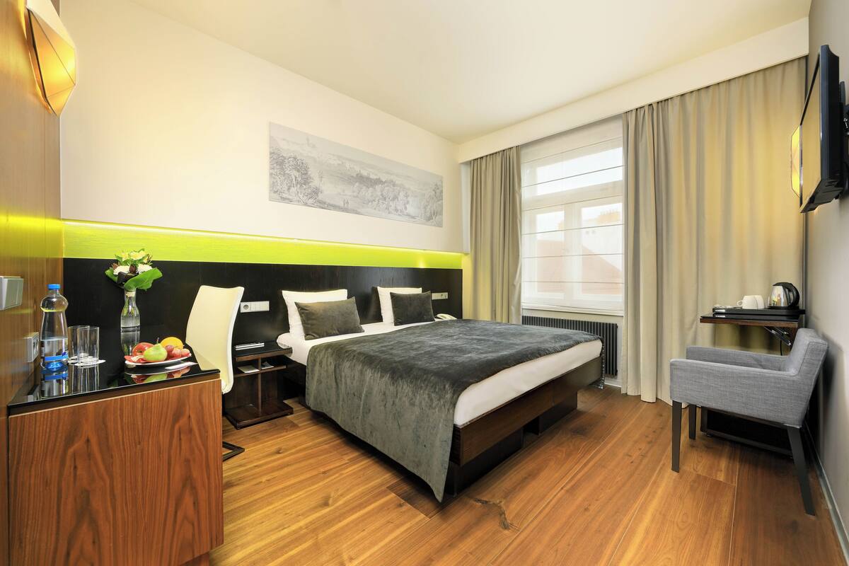 A hotel room with a bed, bedside table, chair, and a window with curtains.
