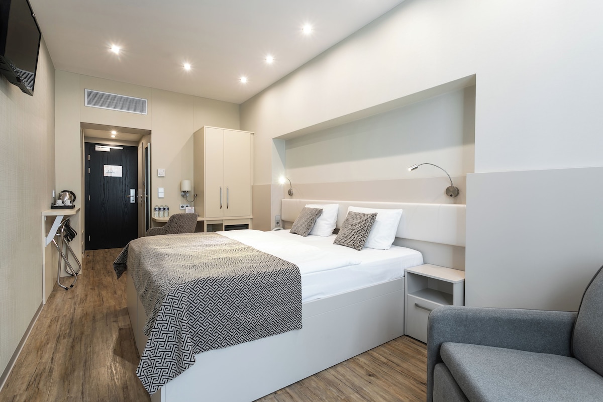 A modern hotel room with a large bed, bedside tables, a chair, and a wardrobe.