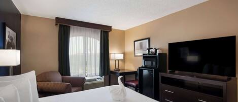 A hotel room with a large bed, a flat-screen TV, a desk with a lamp, a microwave, and a chair.