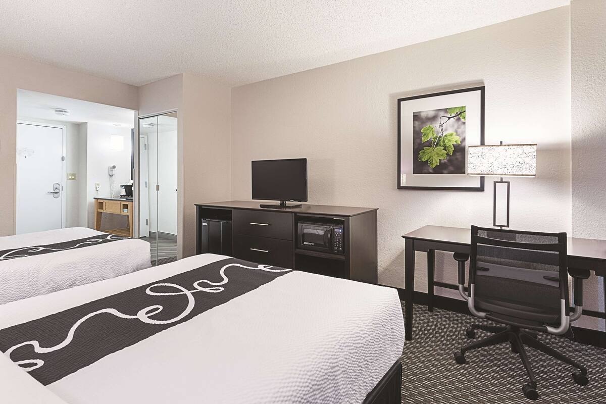 A hotel room with two beds, a desk with a chair, a television, a framed picture on the wall, and a lamp.