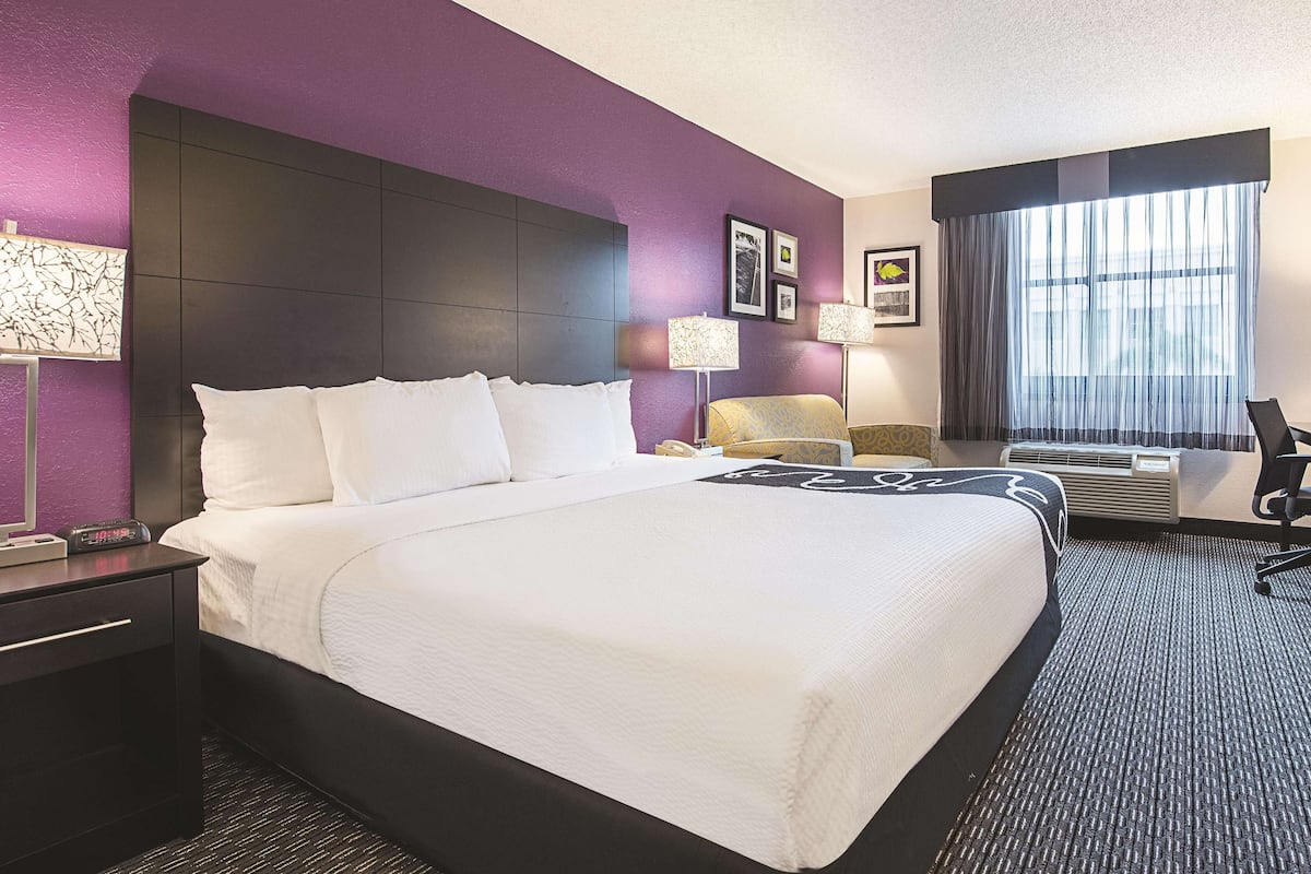 A hotel room with a large bed, a nightstand, a desk, a chair, a desk lamp, a wall-mounted air conditioner, and a window with curtains.