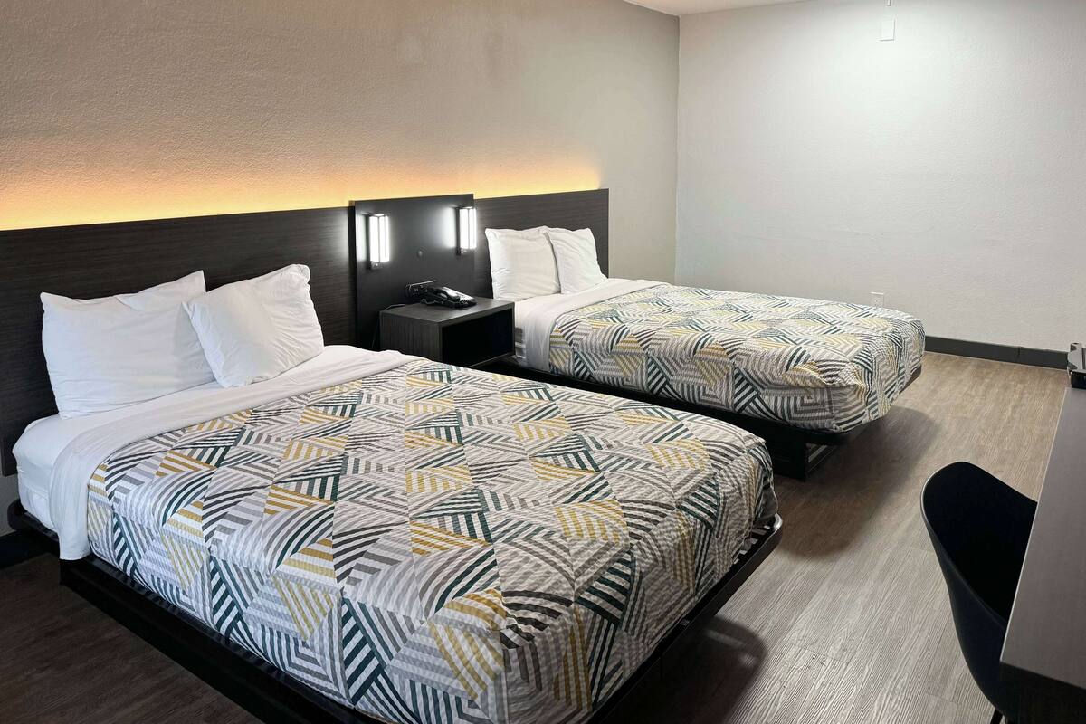 Hotellihuone, jossa on kaksi sänkyä, molemmissa kuviolliset vuodevaatteet, yöpöytä, jossa on puhelin, ja seinälle asennettu valaisin.