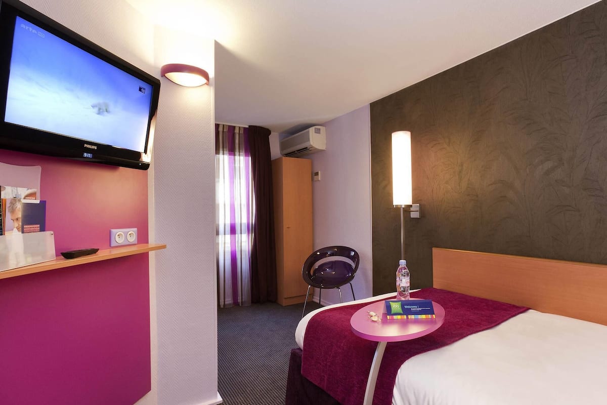 A hotel room with a television, a desk with a remote control, a bed with a red blanket, a nightstand with a bottle and a welcome card, and a chair.