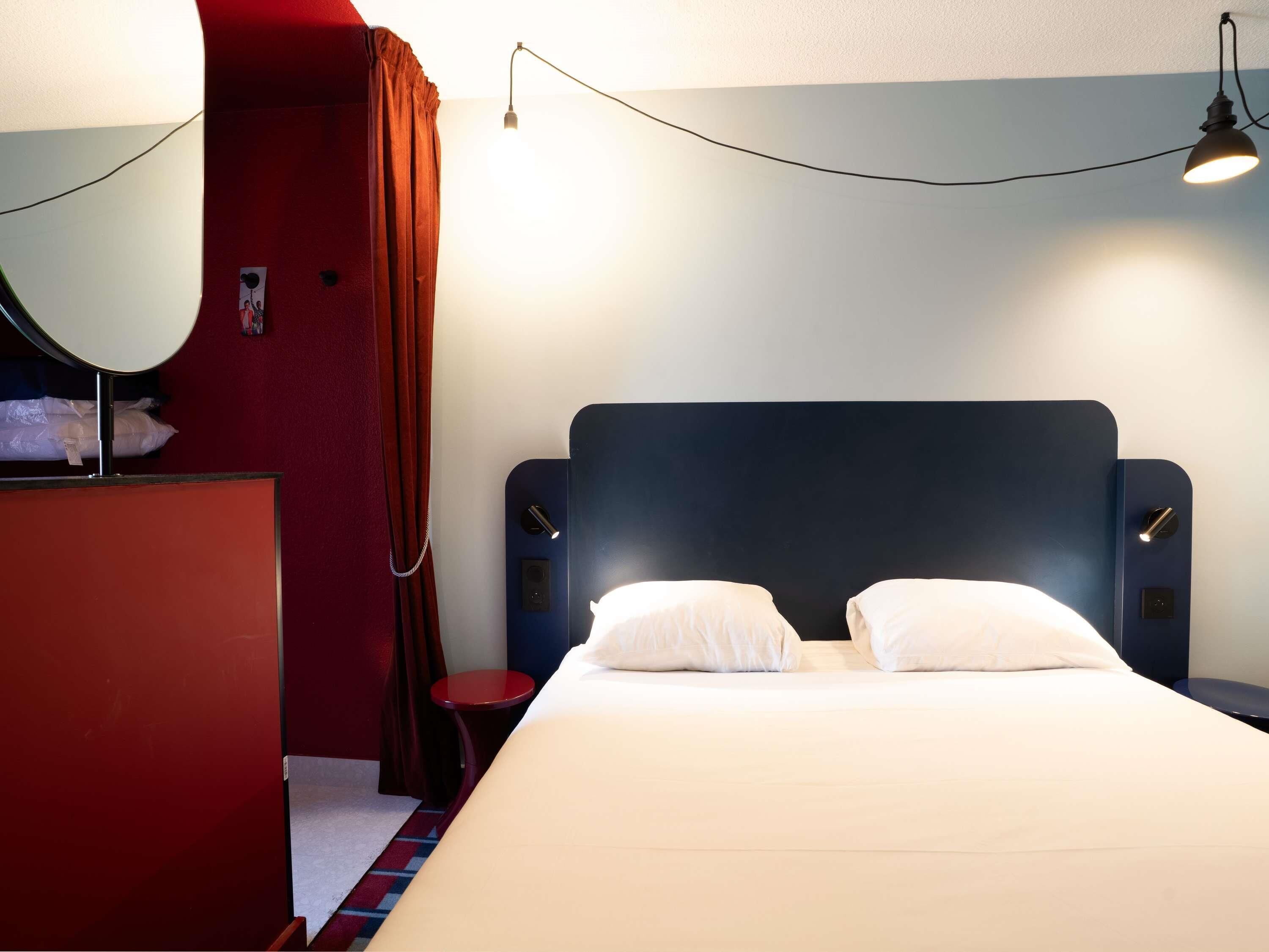A hotel room with a bed, bedside tables, a desk with a mirror, and a red wall.