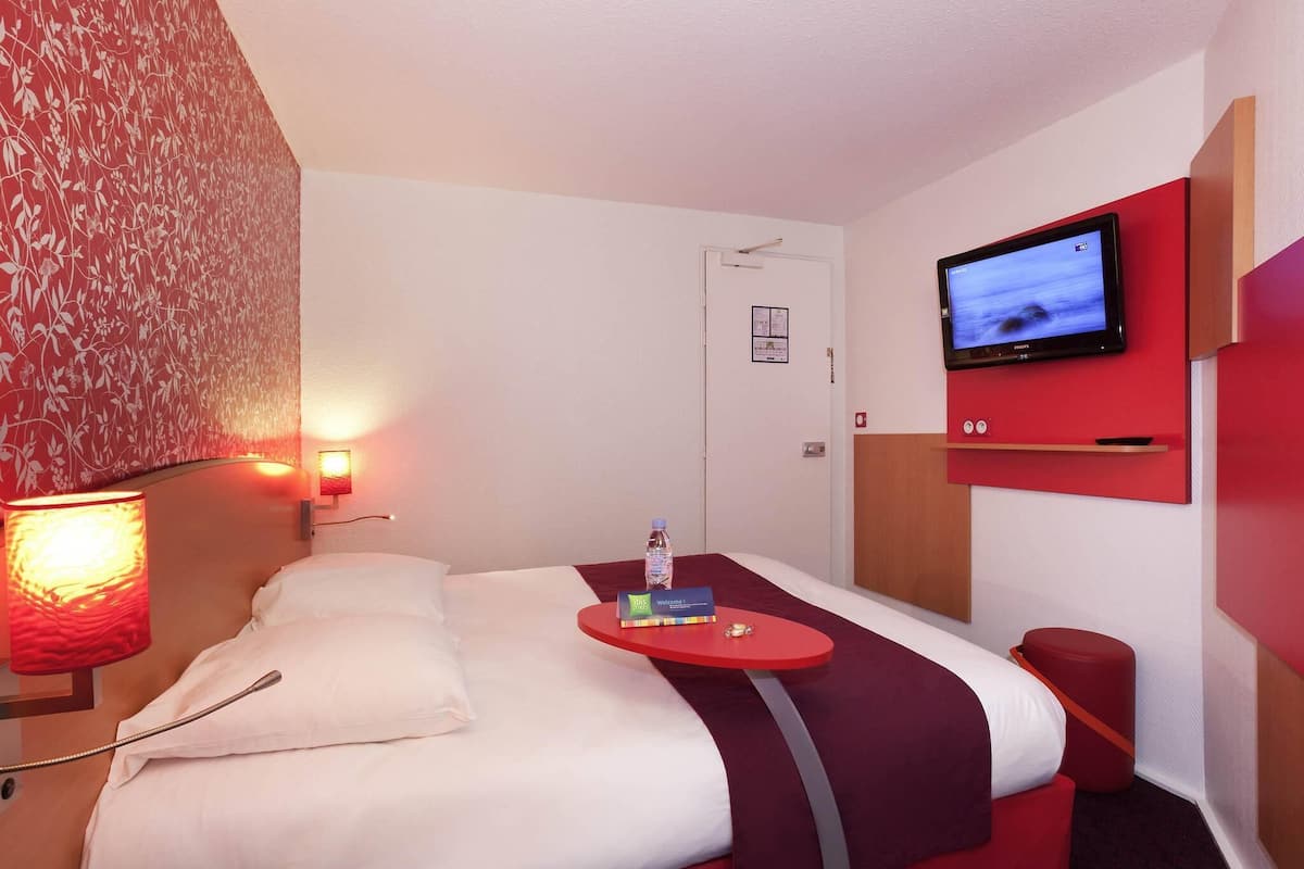 A hotel room with a bed, a TV mounted on the wall, a bedside table with a bottle and a book, and a red accent wall.