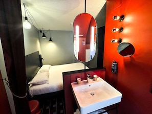 Quarto de hotel compacto com cama, pia branca, espelho e parede decorada em vermelho.