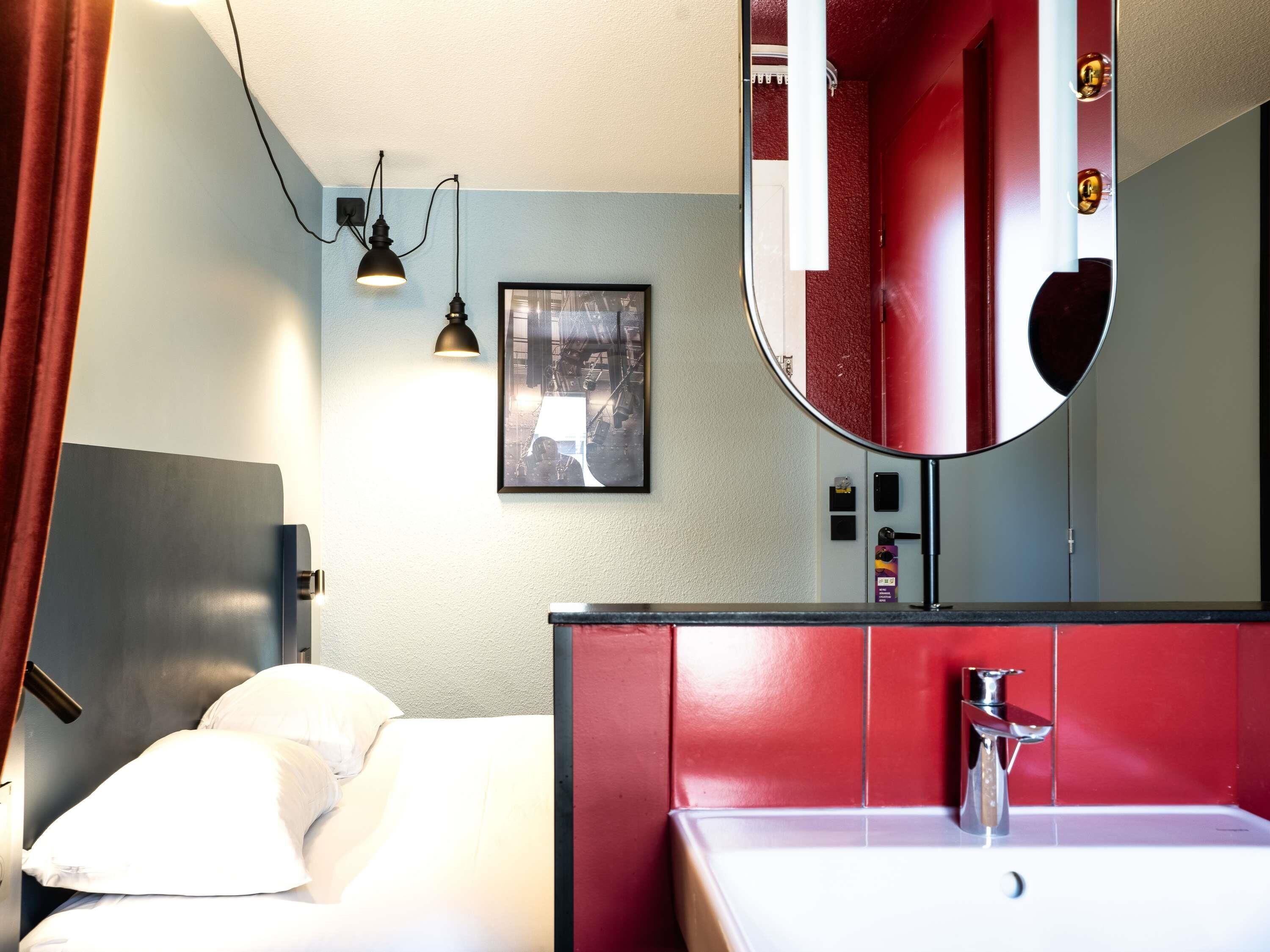 Habitación de hotel con cama, pared de acento roja, espejo grande, lavamanos y un cuadro en la pared.