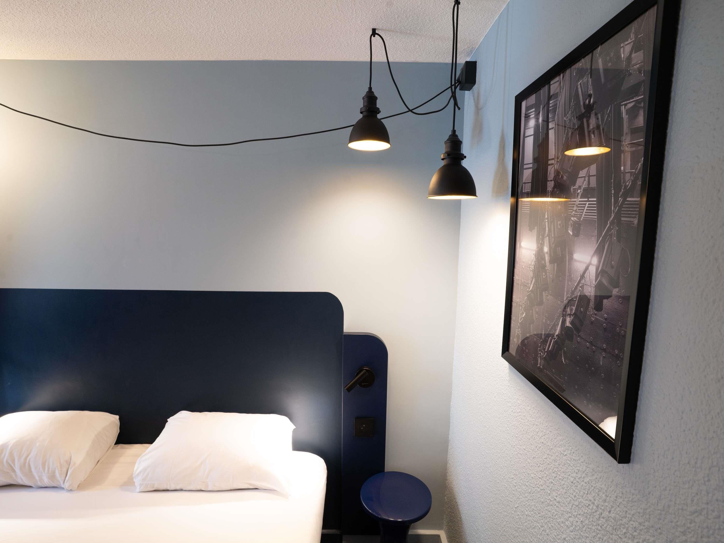 Quarto de hotel com cama, duas luminárias de cabeceira, um quadro na parede e uma pequena mesa redonda.