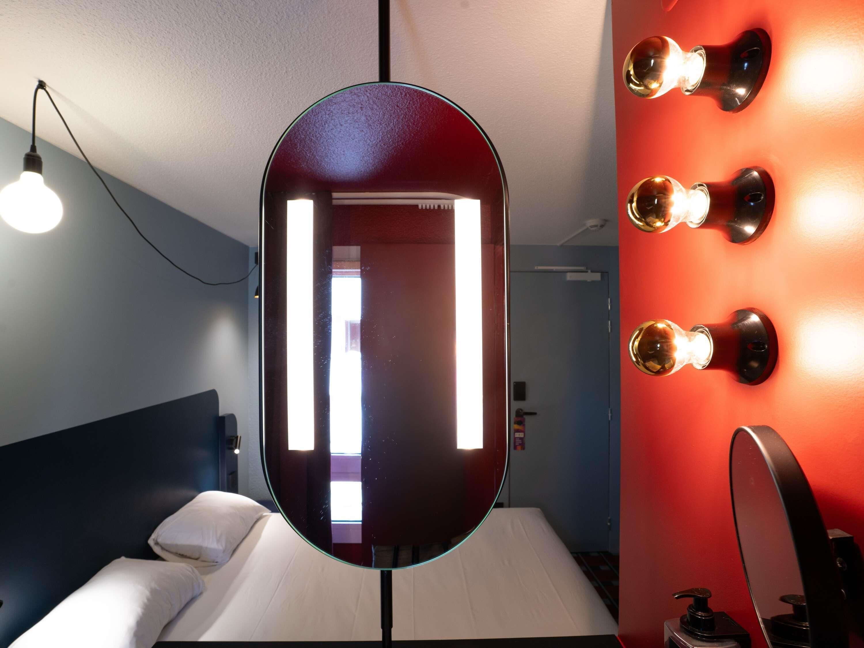 A hotel room with a bed, a large mirror, bedside lamps, and a red accent wall.