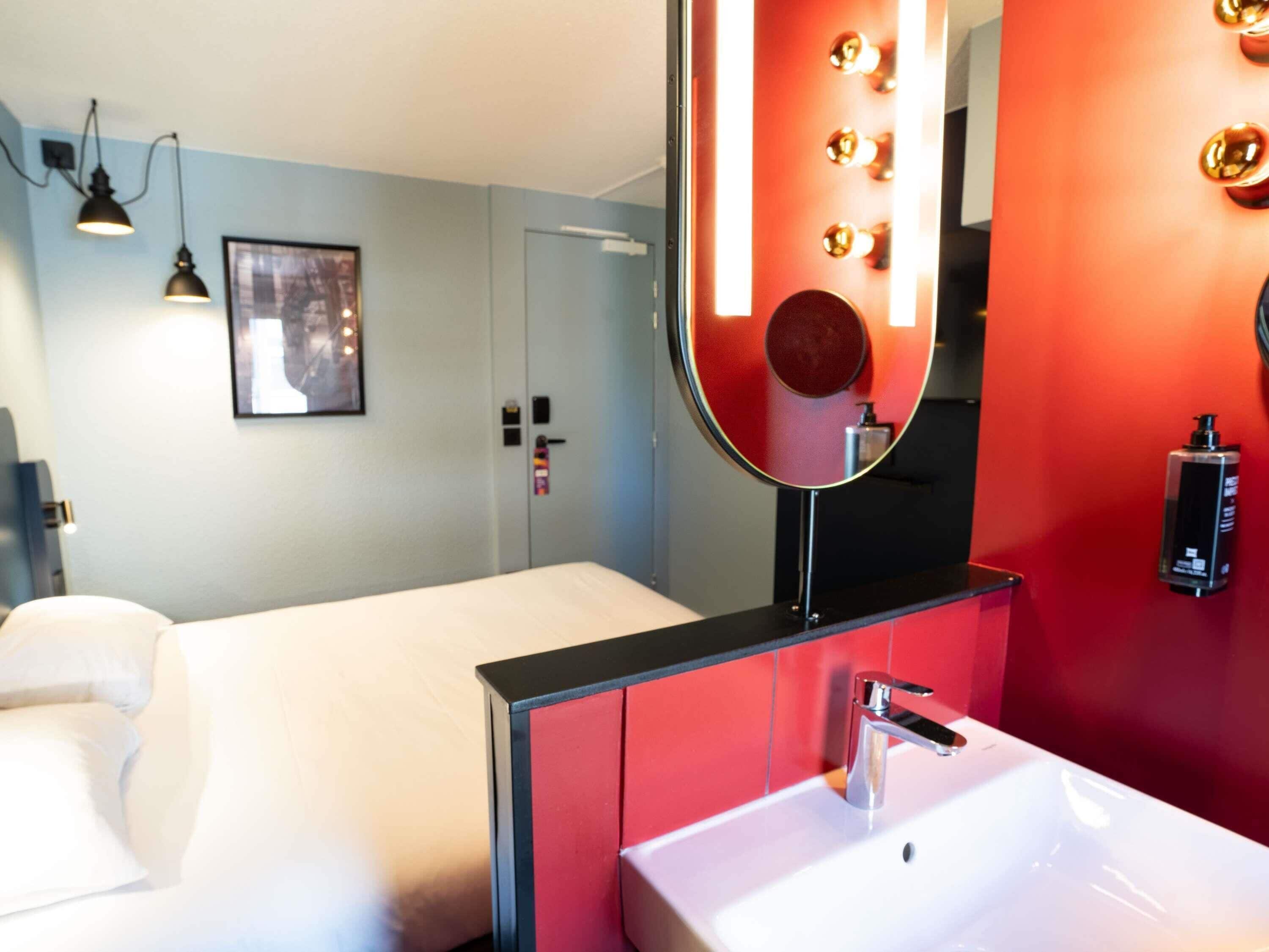 A hotel room with a bed, a large mirror, a sink, and a red accent wall.