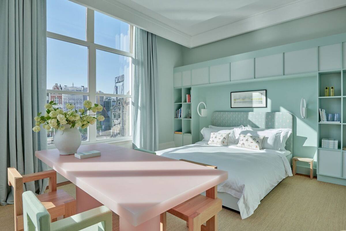 A modern bedroom with a large window, a bed with white bedding, a bedside table, and a bookshelf.