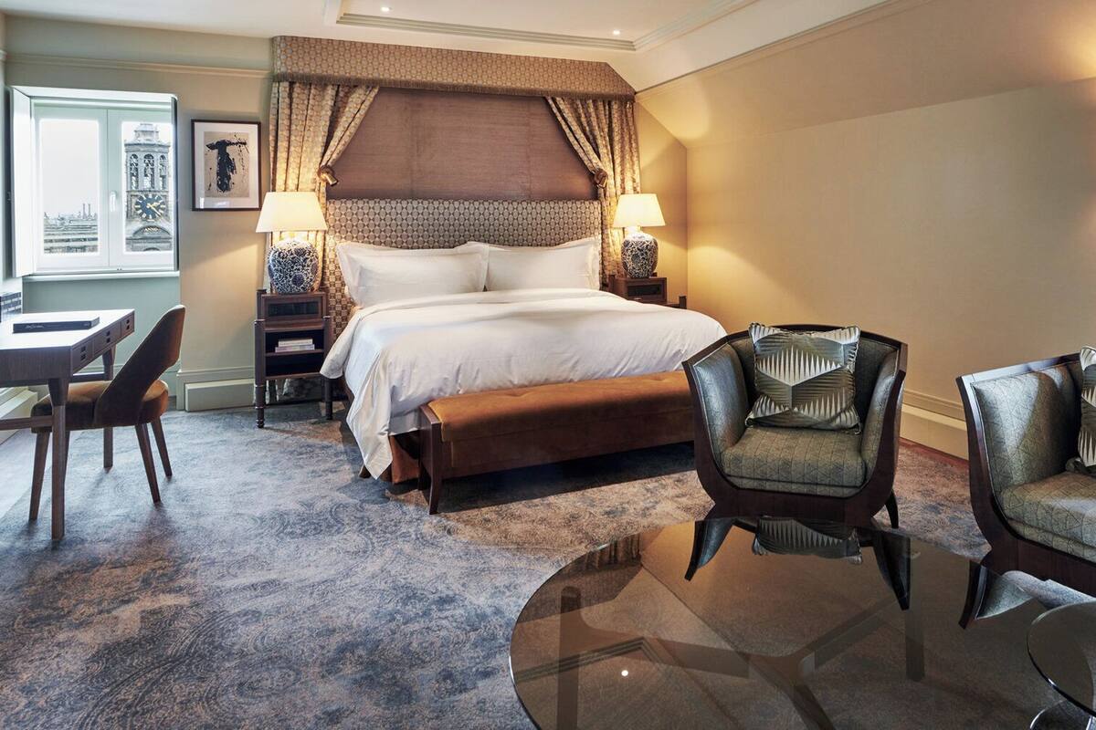A hotel room with a large bed, two armchairs, a desk, and a view of a clock tower through a window.