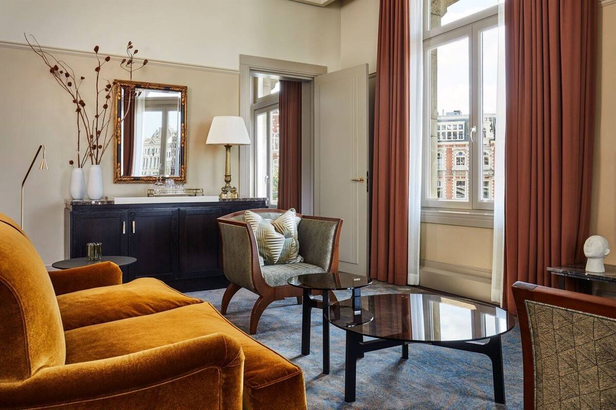 A hotel room with a sofa, a chair, a glass coffee table, a lamp, a mirror, and a view of the city through large windows.