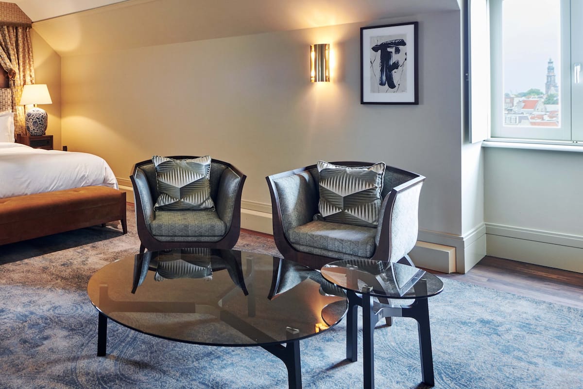 Een hotelkamer met een bed, twee fauteuils, een glazen salontafel, een lamp en een lijst met een kunstwerk aan de muur.
