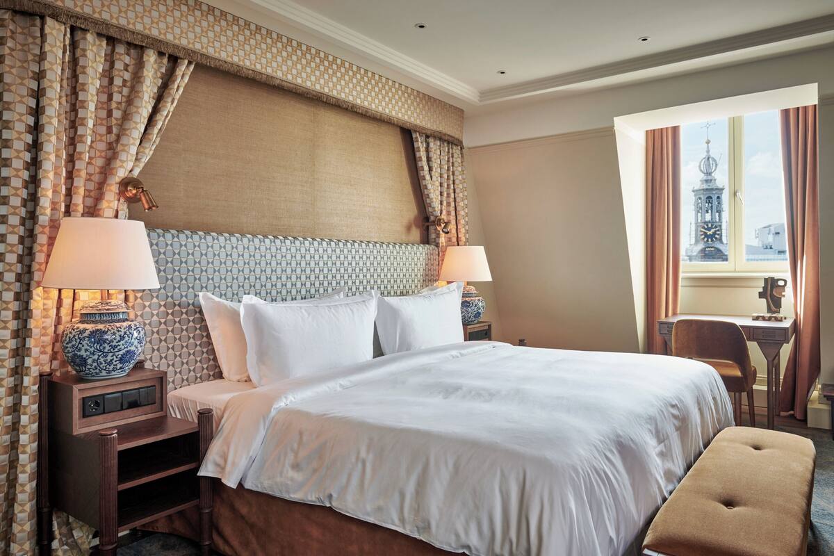 A hotel room with a large bed, two bedside tables with lamps, a small desk, and a view of a tower through a window.
