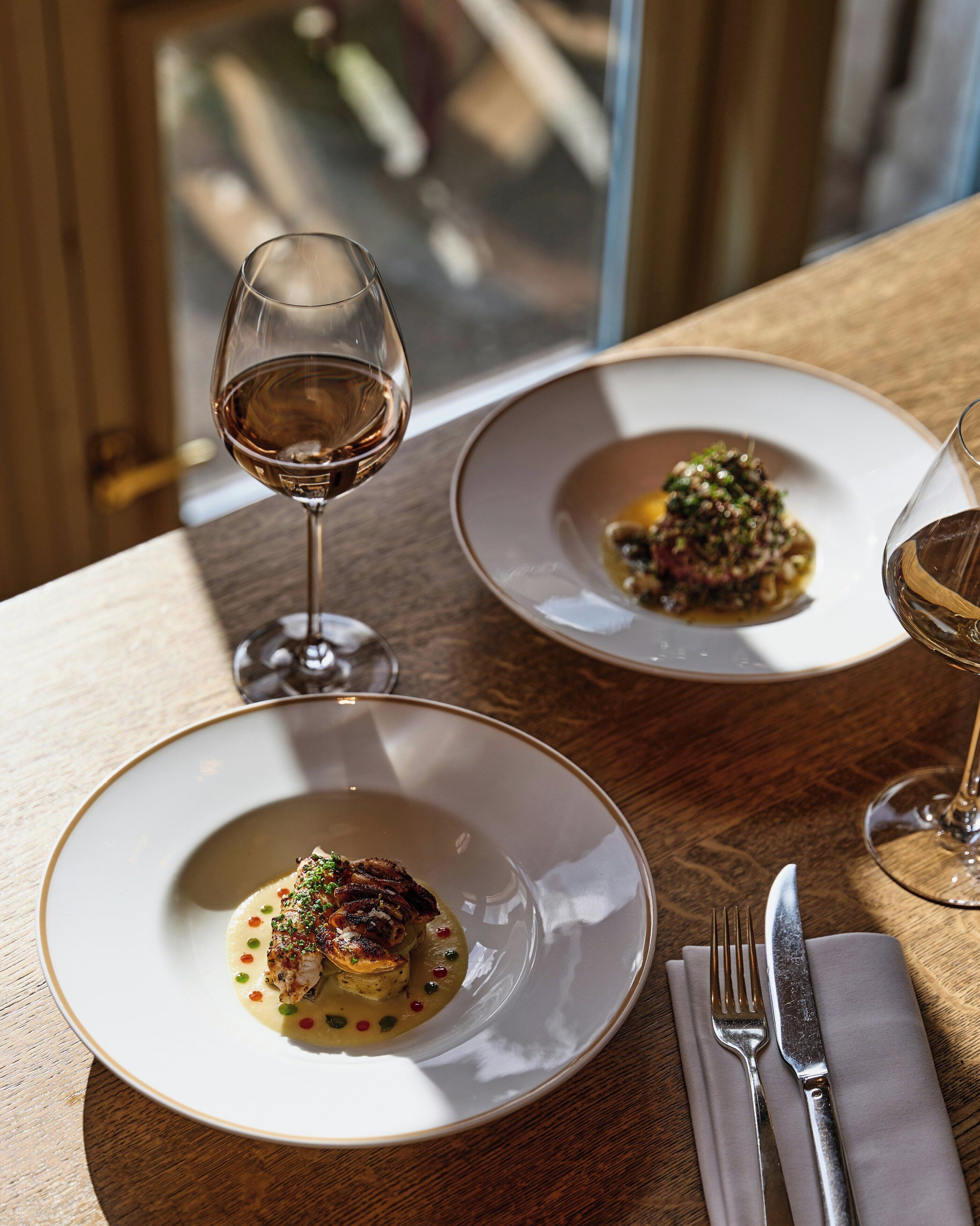 Two plates of food on a wooden table with wine glasses and cutlery.