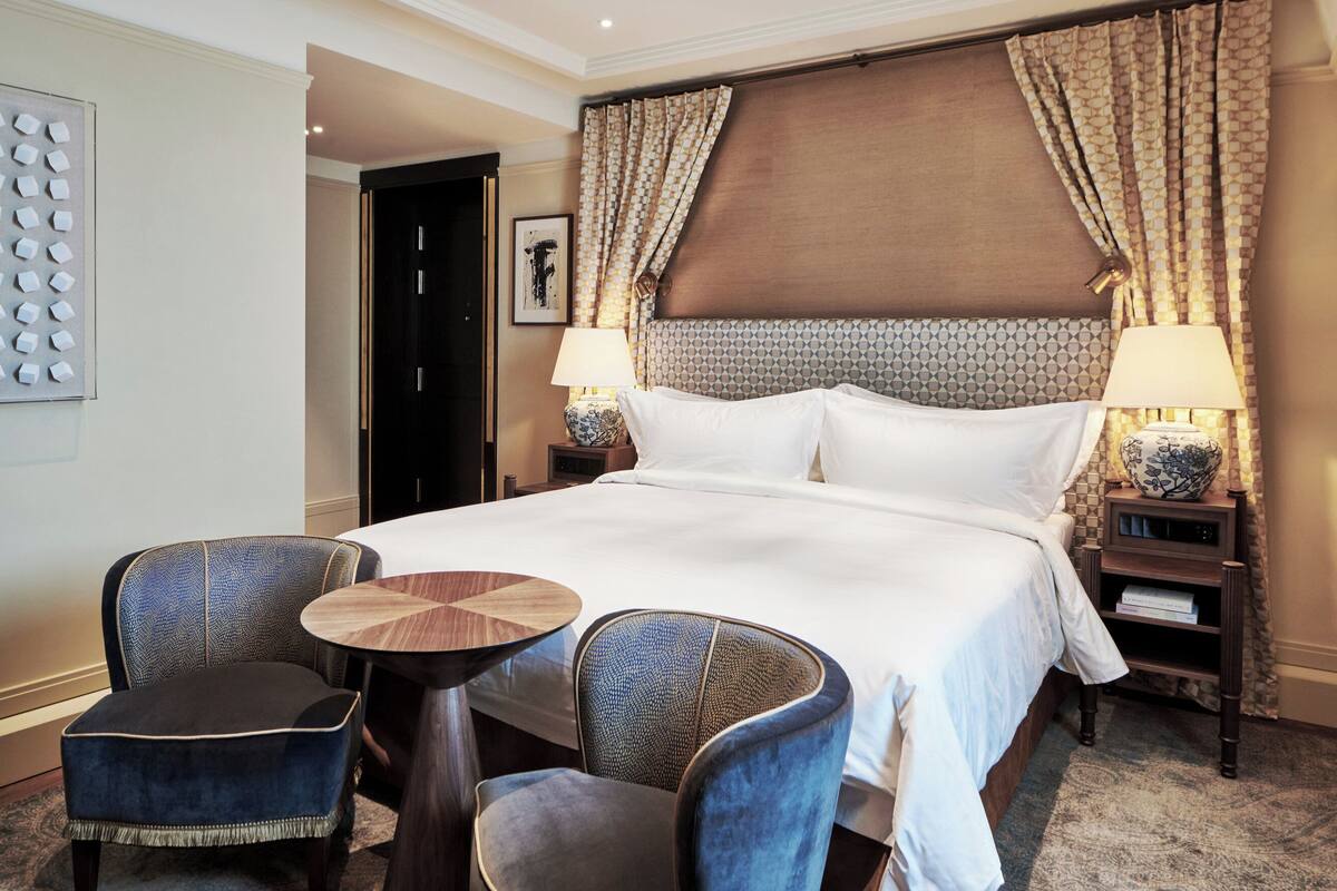 A hotel room with a large bed, two chairs, a small table, a nightstand with lamps, and a decorative wall panel.