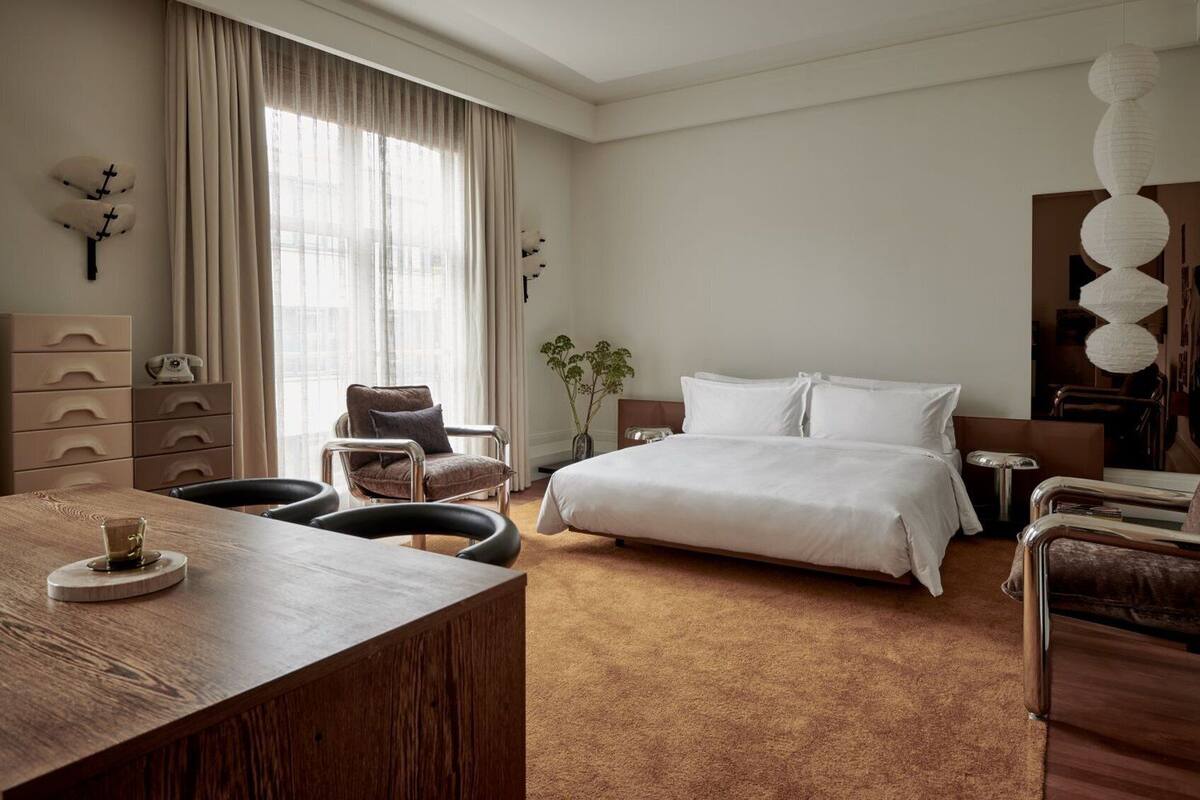 A hotel room with a large bed, a wooden desk, a chair, a small table, and a plant.