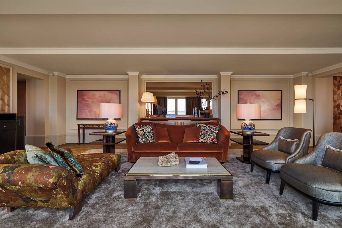 A spacious living room with a brown sofa, a grey armchair, and a coffee table. There are two lamps on side tables and a large painting on the wall.
