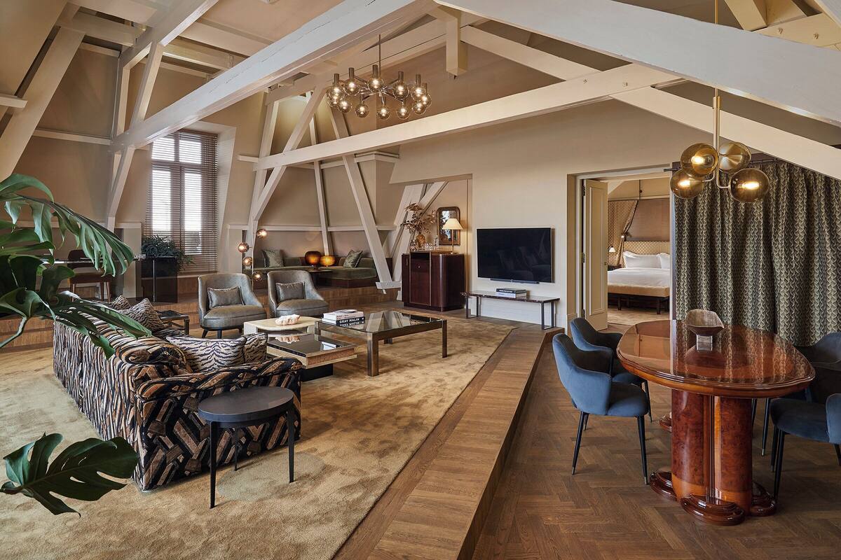 A spacious loft-style living room with a high ceiling, exposed beams, a large sofa, a coffee table, a dining table, and a TV.
