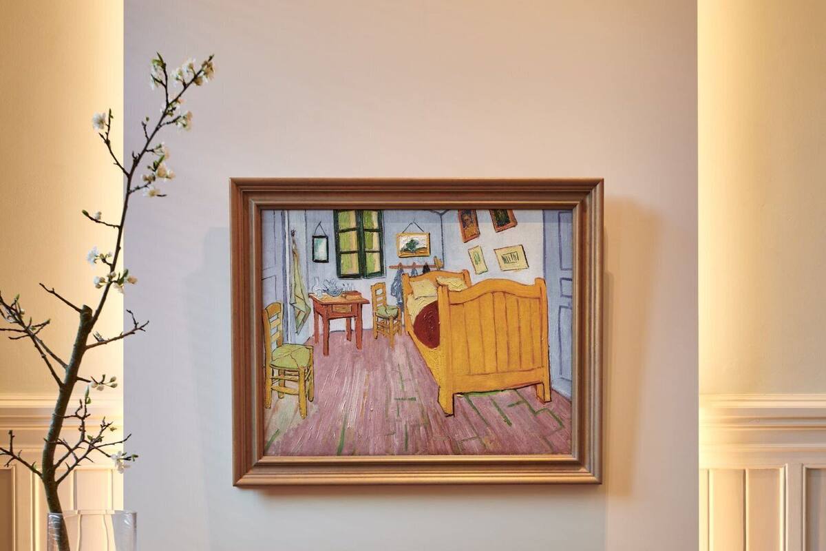 A dining area with a table, chairs, a vase with branches, a framed painting, and a lamp.