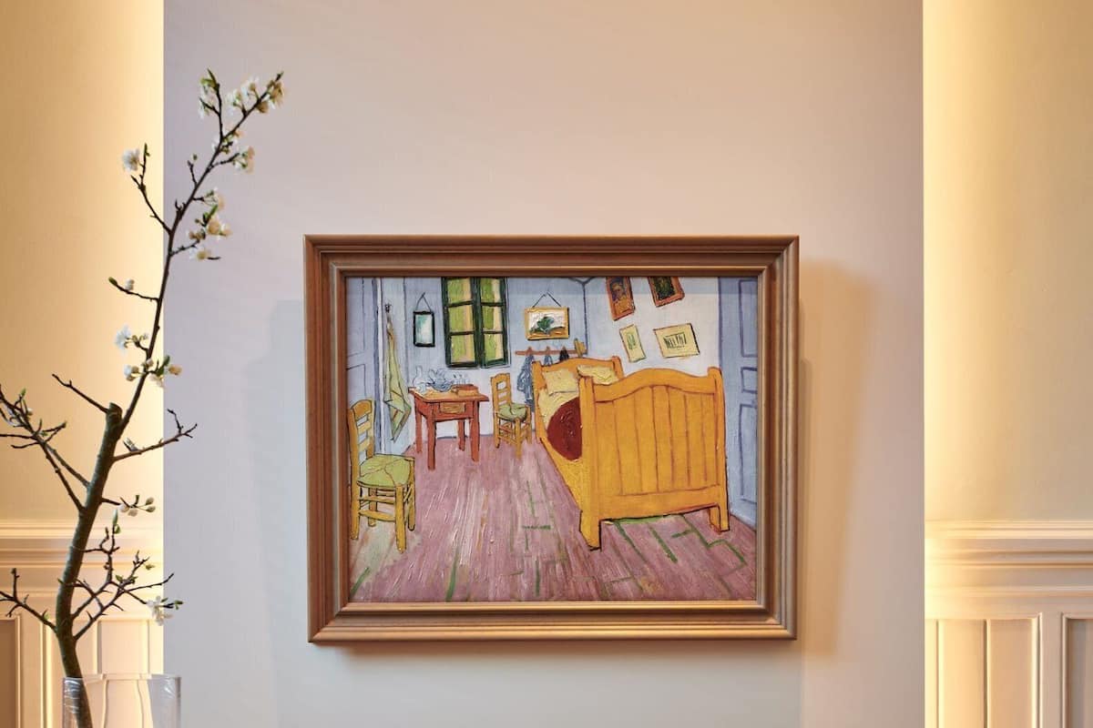 A dining area with a table, chairs, a vase with branches, a framed painting, and a lamp.