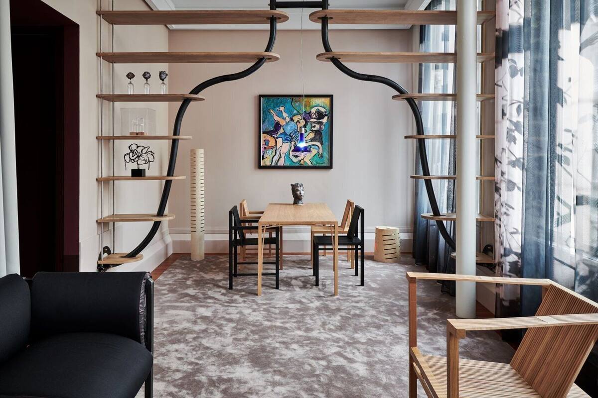 A modern living room with a wooden dining table, black and wooden chairs, a black sofa, and a wooden chair. There is a painting on the wall and a window with curtains.