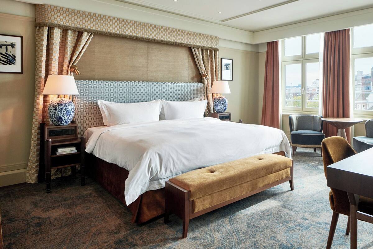 A hotel room with a large bed, a desk, and a chair. There are two lamps on the nightstands, a window with curtains, and a city view.