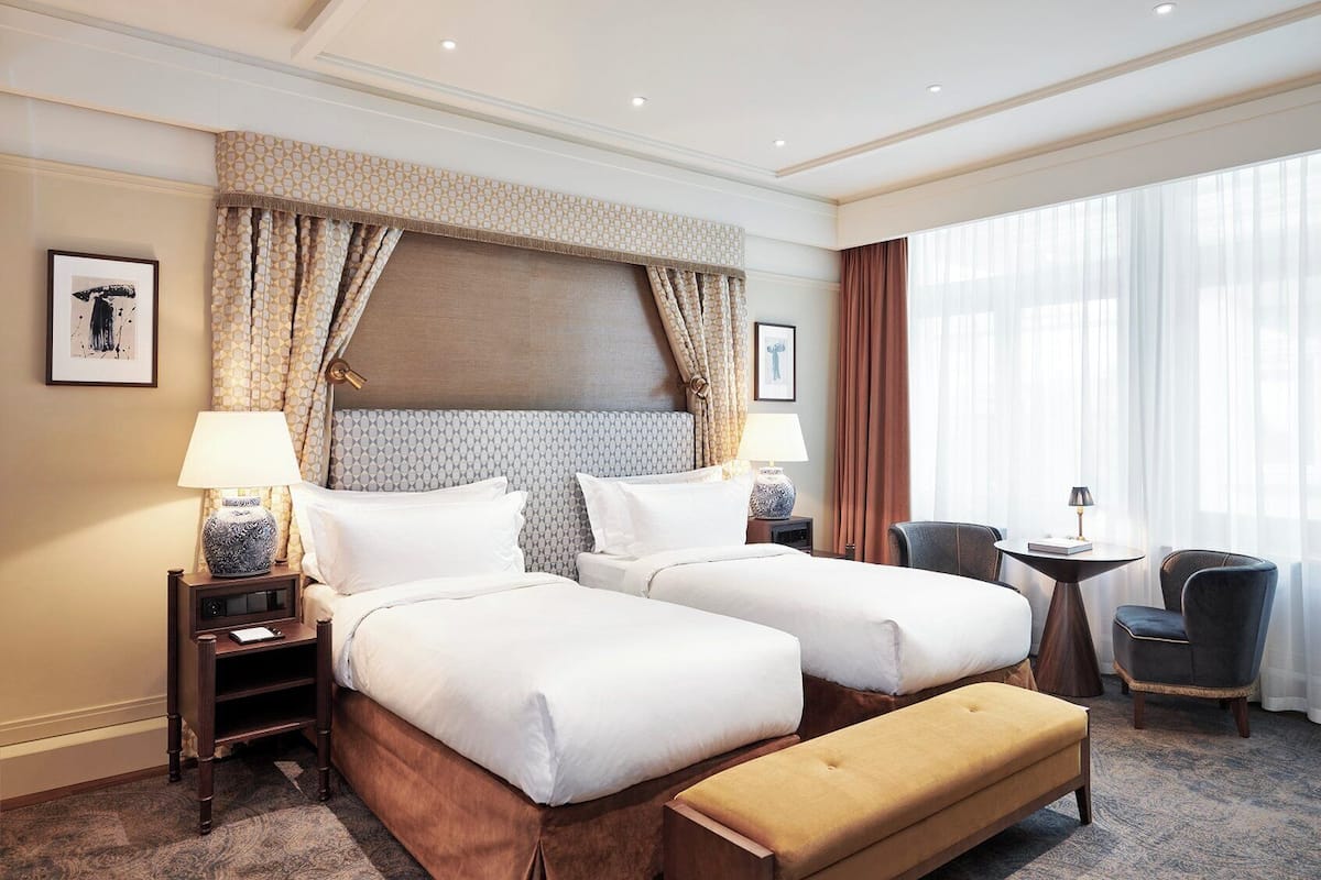 A hotel room with two beds, a bench, a small table, and a chair.