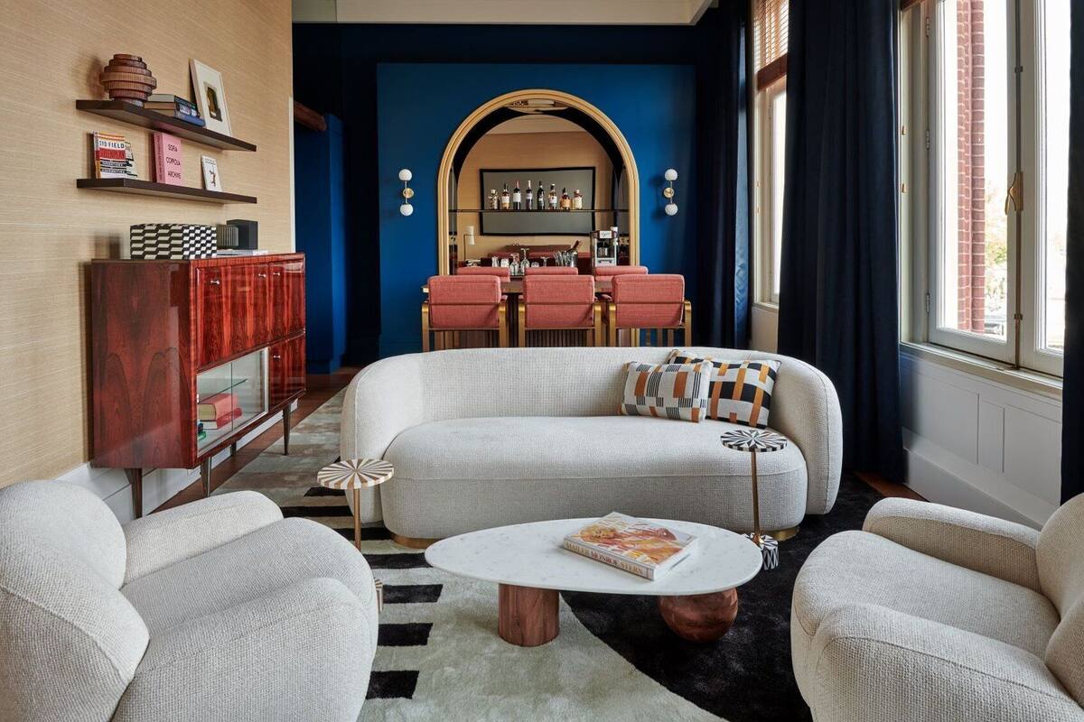 A modern living room with a sofa, armchairs, a coffee table, a bookshelf, and a dining area with a red table and chairs.